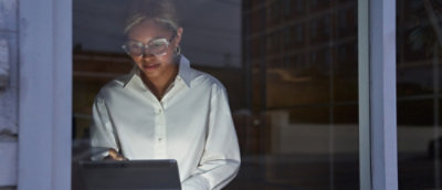 A person wearing glasses and a white shirt stands indoors at night, looking at a tablet or laptop. 