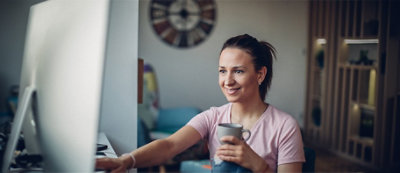 Una donna seduta alla scrivania ha in una mano una tazza e con l'altra usa il grande monitor del computer