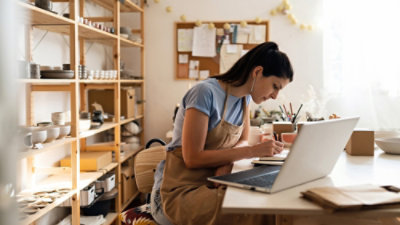  Person in a pottery studio using a laptop and writing on a notepad at a desk.