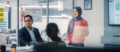 Three people collaborating in a meeting room. One standing, wearing a ping jumper and headscarf, two people sitting at a table. Meeting room with glass walls.