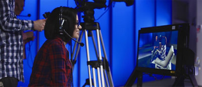 Una persona indossa cuffie seduta davanti a un monitor con una videocamera su treppiedi in una stanza con luce blu.