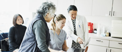 A doctor and medical assistant meeting with two patients in a visit room reviewing medical information on a laptop.