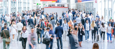 A large transportation corridor with dividers in one section and floor to ceiling windows on both sides. Many busy people are moving in both directions wearing lanyards, carrying work bags and on their phones.