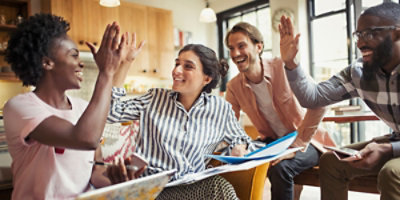 ארבעה אנשים יושבים ליד שולחן, מחייכים ומחלקים "כיפים" זה עם זה, עם ספרים וניירות מולם.