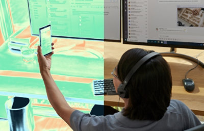 Person wearing headphones working at a desk with two monitors and holding a smartphone.