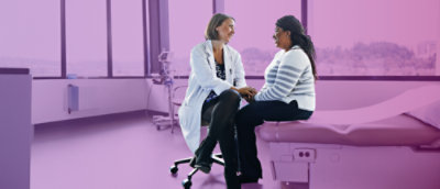 Une femme médecin sur un tabouret face à une patiente assise sur une table de la salle d'examen. Ils se tiennent la main et sourient en parlant.