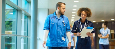 People wearing medical scrubs walk through a hospital corridor while reviewing paperwork and clipboards.