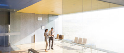  A woman and a man standing in the lobby area of a business building looking at a phone and a tablet PC. The room has floor to ceiling windows on one side and row of benches. 