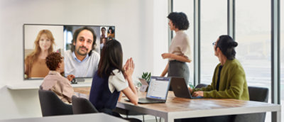 Four people in a meeting room, three sitting down and one standing up, looking at a large screen on the wall with two more people on a video call. Surface device are on the table.