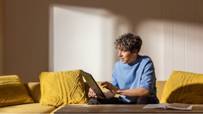 Person sitting on a yellow sofa using a tablet, with sunlight casting shadows on the wall and cushions.
