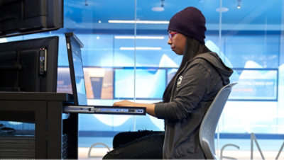 Person wearing a beanie and hoodie seated at a desk, typing on a laptop with an additional monitor, in a modern office setting with blue-toned glass panels in the background.