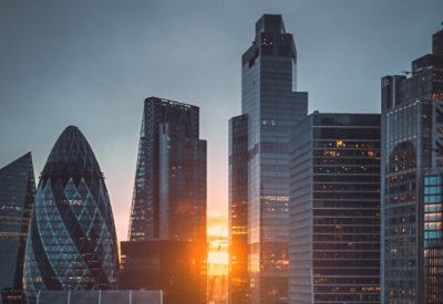 A group of tall buildings in a city with the sun setting behind them.