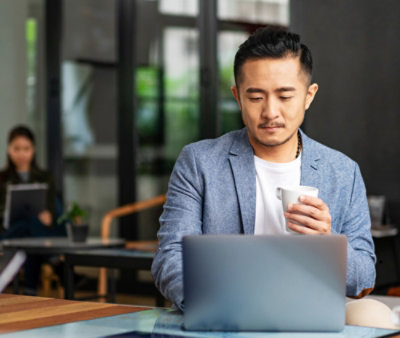  A man holding a cup and looking at a laptop.