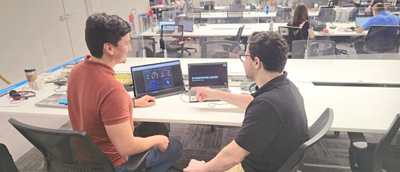 Dois funcionários sorrindo e colaborando enquanto trabalham em laptops em um espaço de escritório moderno e iluminado.