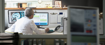 A person with their back to the audience, sitting at a desk and looking at computer screens. One screen displaying images of a brain, another showing data charts.