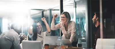 ארבעה עובדי משרד חוגגים סביב שולחן חדר ישיבות עם מחשבים נישאים וניירות, מפגינים התרגשות כשזרועותיהם מורמות באוויר.