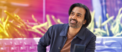 A person, smiling, wearing a dark blue shirt jacket, sitting in front of a flower bed with green plants.