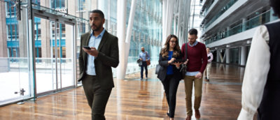 Several people walking inside an open space office building, along a glass façade. One person holding a mobile phone, two other people looking at a table device