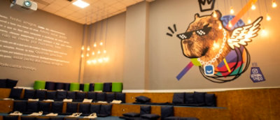 Sala de convivência moderna com almofadas azuis dispostas em arquibancadas de madeira. Na parede, há uma grande ilustração de uma capivara com óculos escuros pixelados, asas e uma coroa, cercado por luzes pendentes. Ao lado, um texto na parede.