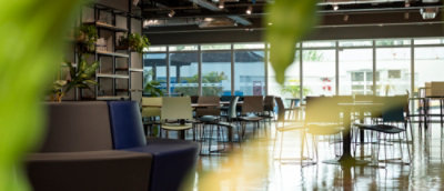 Espaço de convivência moderno e bem iluminado, com várias mesas e cadeiras dispostas de forma organizada. Há plantas decorativas e uma estante ao fundo. Grandes janelas permitem a entrada de luz natural, iluminando o ambiente.