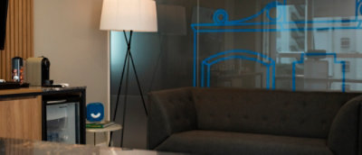 Sala de estar moderna e bem decorada. Há um sofá cinza escuro, uma luminária de chão com um abajur branco, e uma pequena mesa lateral branca com um objeto azul em cima. Ao fundo, há uma parede de vidro com um desenho azul. À esquerda, há um móvel de madeir