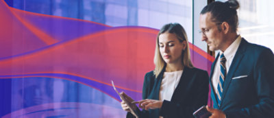  A man and a woman standing to the right of the image, both wearing professional dress. They are looking at a tablet the woman is holding, and the man is holding a cell phone. The background is a blurred window with a red and purple wave floating across.