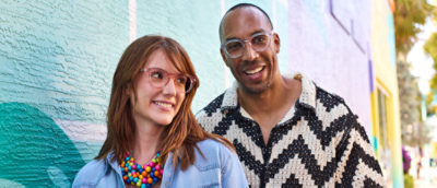 A man of color wearing clear frame glasses standing behind a white woman wearing light pink framed glasses. They are leaning up against a teal blue painted building and both are smiling.