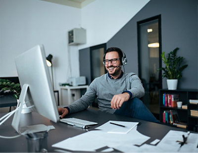 A man wearing a headset, seated at a desk, smiles while looking at a computer screen in a modern office setting.