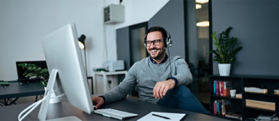 A man wearing a headset sits at a desk with a computer, smiling and looking at the screen
