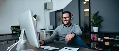 A man wearing a headset sits at a desk with a computer, smiling and looking at the screen