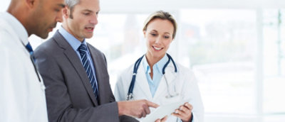Three professionals, two in white coats and one in a suit, discuss a document.