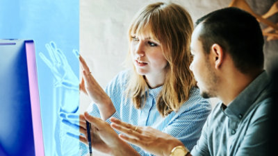 Two people in discussion, gesturing toward a computer screen displaying a blue-toned graphic