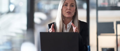 Person seated at a desk, gesturing with hands while working on a laptop in a modern office environment