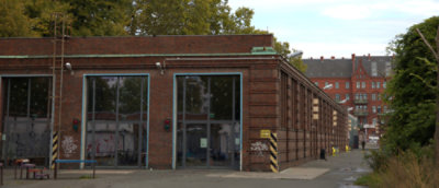 The image depicts a brick industrial building with large windows and a ladder on the left, a "Schritt fahren" sign on the right, and another red brick building in the background.