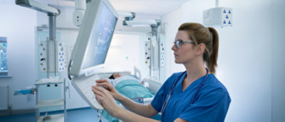 A person in blue surgical scrubs wearing a stethoscope looking at a monitor with a patient lying on a bed in the background.