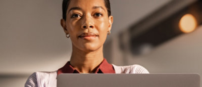 A confident woman with short hair looks at the camera over a laptop, wearing a red blouse and a light-colored sweater.