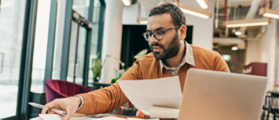A person sitting holding pieces of paper with a laptop on the foreground.