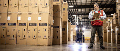Warehouse worker wearing a safety vest holds a clipboard while inspecting stacks of large boxed appliances, with a forklift operating in the background.