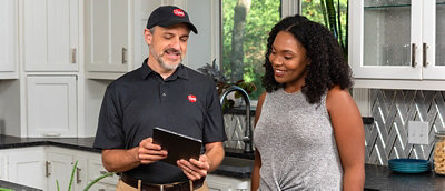 The image shows a modern kitchen with white cabinets, a black countertop, and a geometric backsplash. A person in a black uniform with a "Rheem" logo and cap holds a tablet, discussing something with another person in a gray top.