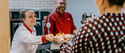 A chef in a red uniform hands a plate of food to a person in a patterned shirt, with another chef in a white uniform assisting.