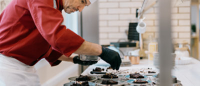 A chef in a red uniform and white apron uses black gloves to place brownies on plates in a modern kitchen with a brick wall and various kitchen equipment in the background.