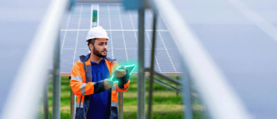 Field technician in safety gear using a digital tablet while inspecting solar panels at a renewable energy installation site.