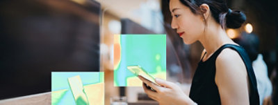 Person holding a smartphone and working on a computer at a wooden desk in a modern indoor setting.