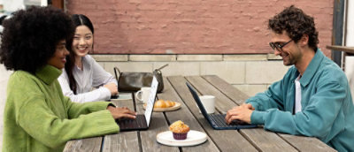  Kolm inimest töötavad sülearvutitega õues puidust laua taga, kus on kohvitassid ja saiakesed.