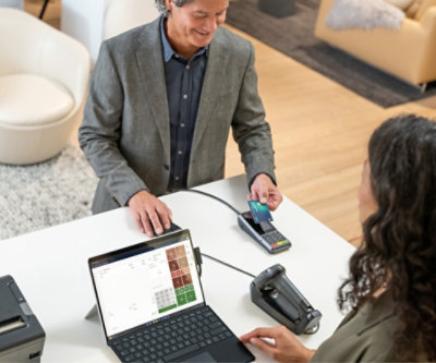 ผู้ชายยืนอยู่ที่จุดเช็คเอาท์ของร้านค้ากําลังยิ้มให้พนักงานเก็บเงินหญิงที่นั่งอยู่ซึ่งกําลังคิดเงินโดยใช้ตัวอ่านบัตร