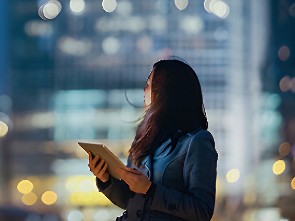 A woman in a business suit holding a tablet looks upward at night, illuminated by city lights