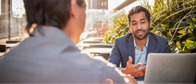 Two individuals engaged in a conversation at an outdoor table with a laptop open in front of them.