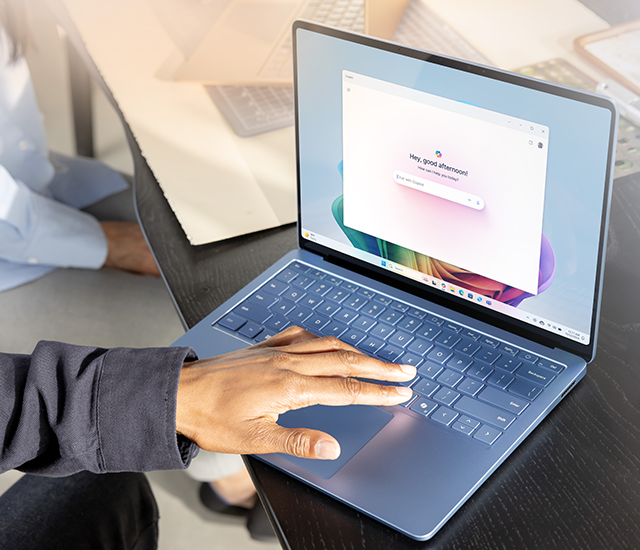 A person wearing a dark sleeve operates a blue laptop on a black wooden desk. The laptop screen shows a browser with a greeting and an input box. Sunlight softly illuminates the scene. Partial views of another person, papers, and a tablet are visible in the background, suggesting a collaborative office environment.