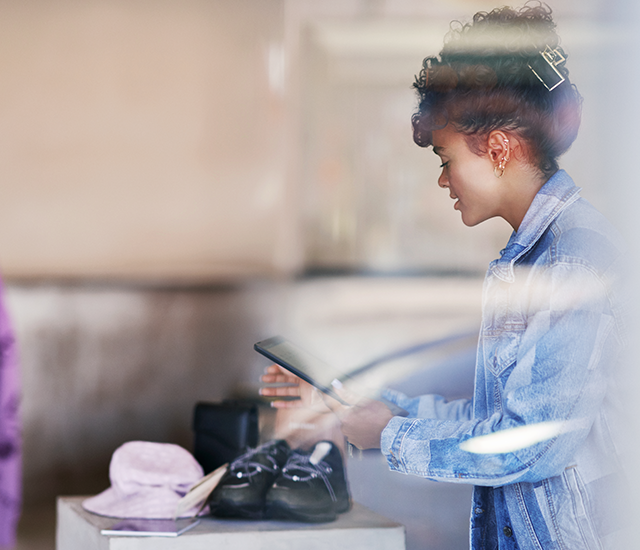 A person in a denim jacket interacts with a tablet in what appears to be a store setting. In the foreground, a soft pink hat and black shoes are on display, suggesting a retail environment. Sunlight streams through a window, creating a casual and professional atmosphere. The scene reflects a blend of technology with traditional shopping experiences..
