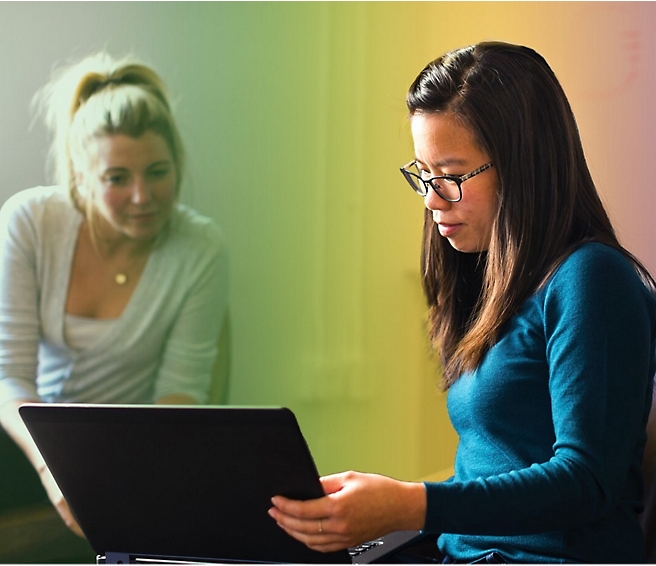 Hai người phụ nữ trong một không gian văn phòng thông thường; một người làm việc trên máy tính xách tay trong khi những người còn lại đang chăm chú xem